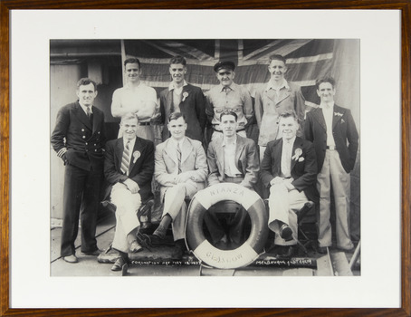 Framed black and white photograph depict the crew of the Nyanza, Glasgow posing and dressed up for the occasion. 6 are standing in front of the Union Jack and 4 are sitting on a bench., the Nyanza buoy at their feet.