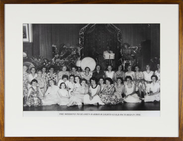 Black and white group photograph of Women from the Harbour Lights Guild sitting on the ground and benches  in front of the stage at the Flinders Street Mission during a musical event.