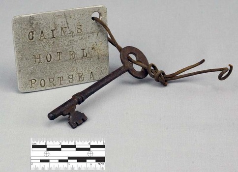 Door key featuring one set of teeth and a rounded handle at the end. Looped through the hole of the handle is a piece of twisted wire attached to a square tag. 