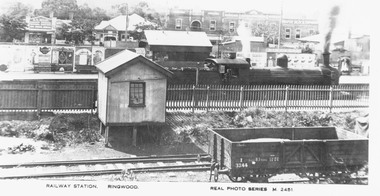 Photograph, Steam locomotive at Ringwood  Railway Station, with platform advertising boards and shops fronting Maroondah Highway.  Photographed looking north from Station Street, Ringwood - Postcard c. 1920"