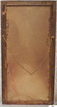 The handwritten inscriptions are sideways along the lower left edge of the painting’s board, red writing left of the pencil writing. The board is held in place inside the timber frame with small metal tacks around all sides. Staples join string to each side of the painting for hanging it. The string is hanging in a downward curve to a point where it would be resting on the hook. Small round nail heads are dispersed around the frame and there is a change of colour on both the board and the wood; perhaps from having a backing paper taped and tacked to the back. There is some damage and watermarks on the board.