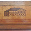 Inlaid wood depicts a building with dark roof and side, a chimney, and a light coloured front. 