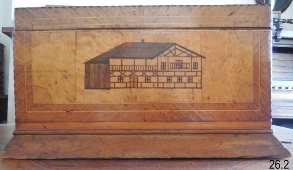 Inlaid wood depicts a building with dark roof and side, a chimney, and a light coloured front. 
