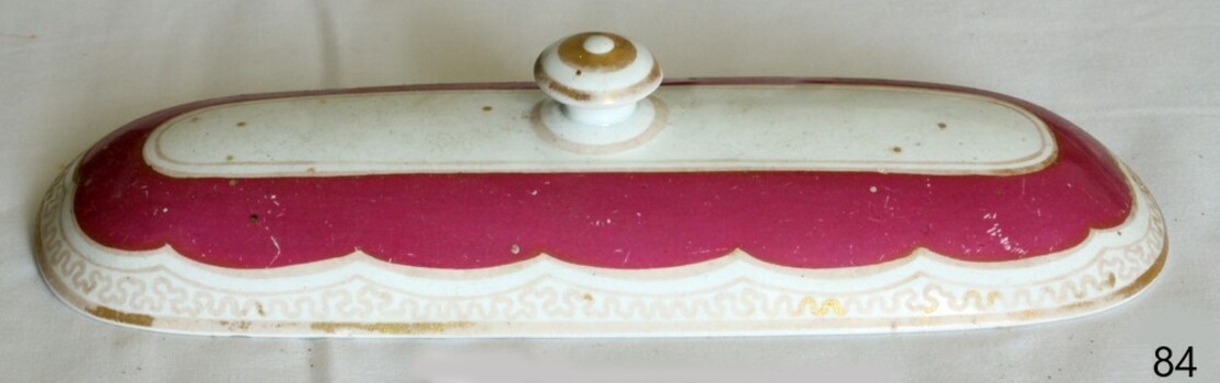 White rectangular ceramic lid has dark pink section around handle and gold highlight pattern around edge