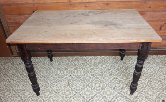 Oblong wooden table on four dark brown legs. The table surface appears to have been scrubbed clean multiple times, resulting in it being reduced to light coloured bare wood.