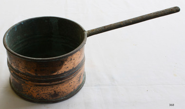 Small copper saucepan with long handle, showing signs of corrosion. Three ridges around the circumference, halfway down the exterior. 