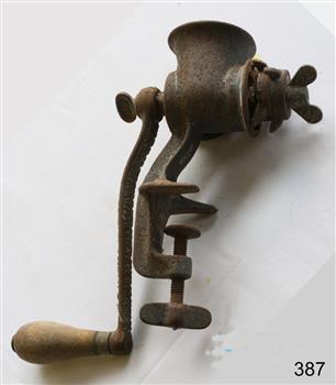 A metal mincer with a handle for rotating the mincing apparatus inside. There is a clamping mechanism to fasten the mincer to the kitchen bench or table. The item is very rusty.