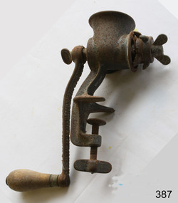 A metal mincer with a handle for rotating the mincing apparatus inside. There is a clamping mechanism to fasten the mincer to the kitchen bench or table. The item is very rusty.