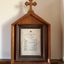 The document contains a list of text. It is behind glass in a timber frame, the top edge is an inverted V with a cross