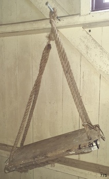 Plank of wood with three reinforcing wooden sections underneath, ropes looped and ends tied below.