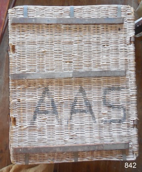 The wicker lid has been strengthened by the addition of wooden braces attached with metal straps above and below the lid. The lid is attached to the basket at the back with strong metal rings. The large black stencilled letters "A A S" can be seen on the lid.