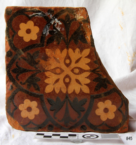 Square brown tile, one corner broken, with black and cream inlaid floral pattern