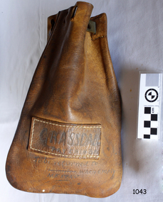 Leather bag, rounded bottom corners, holes in top for drawing together, stitched frame around metal nameplate