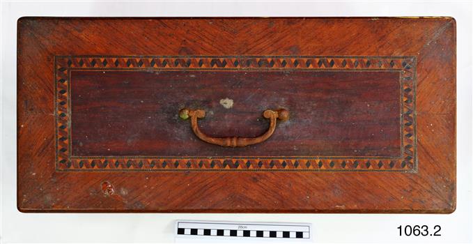 The lid is made from four pieces of timber , each turned around so that the grain runs at an angle towards the centre. In the centre is inlaid a darker rectangle of timber with the metal handle  in the centre of it. In between the outer timbers and the centre timber panel is a rectangular patter of different colours.