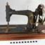 The front of the sewing machine is facing the viewer. Gold decals decorate the metal work. The crank handle is on the right hand side of the machine. Below the needle, on the bed of the machine, are three metal plates. The plate on the right side of the needle has the maker's details and the serial number.