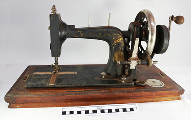 The front of the sewing machine is facing the viewer. Gold decals decorate the metal work. The crank handle is on the right hand side of the machine. Below the needle, on the bed of the machine, are three metal plates. The plate on the right side of the needle has the maker's details and the serial number.