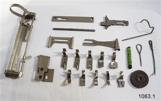 The set of accessories is laid out on the table. Most of the parts are a variety of feet to use with the machine for different effects and purposes. There are also some metal tools, a marking crayon and other parts.