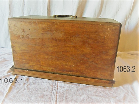 The wooden case is clipped onto the base of the machine, leaving the bottom of the base still visible. It has a folding metal carrying handle on the top. 