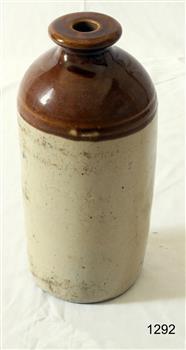 Bottle has wide lip around mouth. It has a shiny brown glaze from top to just below the shoulders. The rest of the bottle is a cream colour. The slightly rough texture of the clay can be seen.