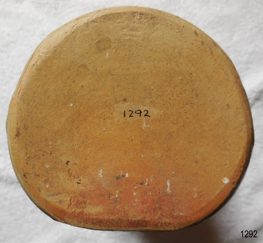 The flat side and arched side of the bottle can be seen from the shape of the base. The unglazed clay looks rough and has pits and marks. There are spots of white, rusty brown and crimson colour in places. The handwritten museum registration number is clearly visible.