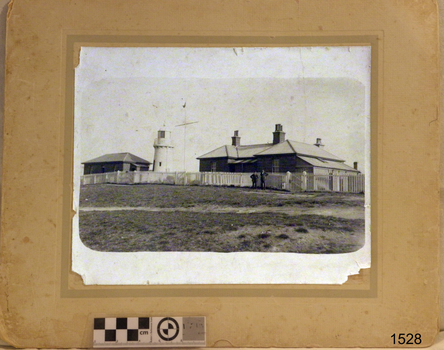Black and white image of a building, lighthouse, flagstaff and cottage with picket fence and gate. Two male figures stand in front.is in the front.