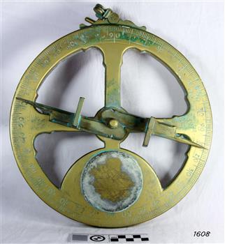 Gold-coloured disc shape with cup-outs, an indicator arrow, and a ring on the top.