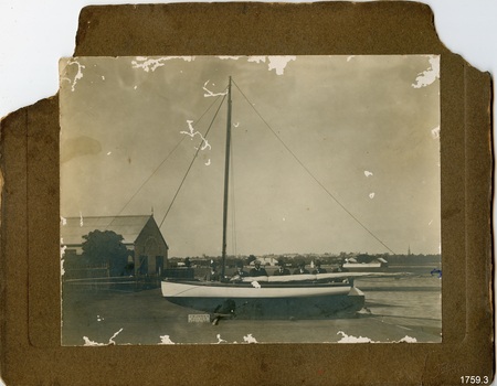 Sepia photograph mounted on card, stamped lower right. Seven men standing behind a small sailing boat.