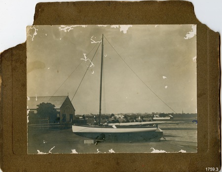 Sepia photograph mounted on card, stamped lower right. Seven men standing behind a small sailing boat.