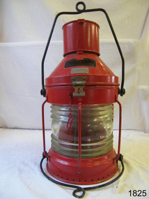 Red metal lamp with glass all around, holes formed all around base, cylinder on top of lid, brass nameplae on side.