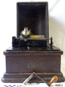 Phonograph with lid off, crank handle on bench in front of machine.