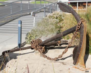 Small anchor on its side with head and fluke on the sand, chain attached.