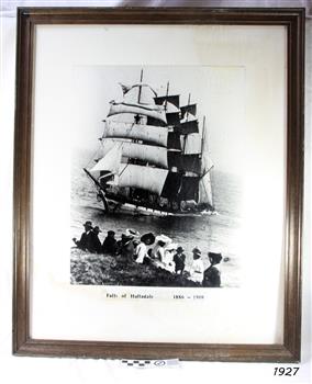 Framed black and white photograph. People seated on the ground, viewing a sailing sip