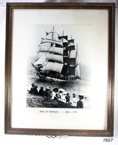 Framed black and white photograph. People seated on the ground, viewing a sailing sip