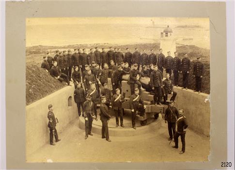 Group of figures in military uniform standing around a cannon that is facing towards Lady Bay