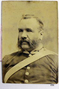 Uniformed male with beard and moustache. Uniform has light coloured buttons and a sash.