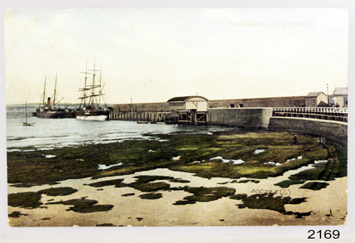Tinted postcard of the Warrnambool Breakwater showing docked vessels, small buildings and the beach.