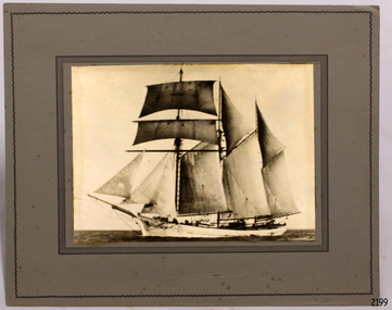 Sepia photograph mounded on card that has a printed border. Subject of ship in full sail