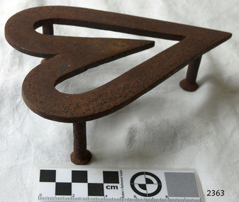 Heart shaped cast iron trivet standing on three supports. The inside area of the trivet is open. The trivet was painted black, but is now mostly rusty.
