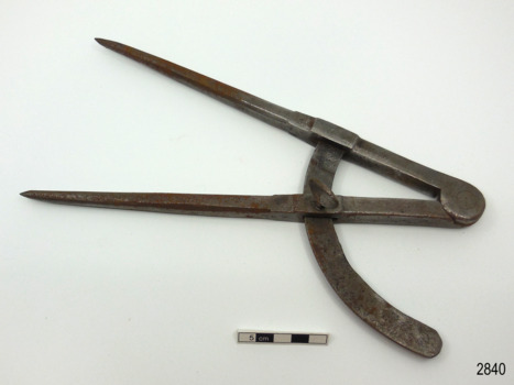 Metal compass has a 90 degree arc and a screw to hold the angle in place. The pointed arms are hinged at the top. There is an inscription on the compass. 