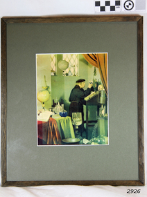 A robed male standing at a desk inside a room with flasks and bottles close-by