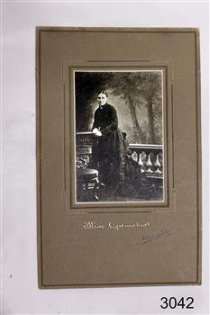 Woman standing in dark Victorian era outfit, short column fence behind, trees in background