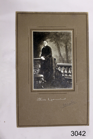 Woman standing in dark Victorian era outfit, short column fence behind, trees in background