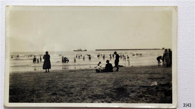 Black and white photograph with shore, water and people bathing. Ship in background.