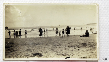 Black and white photograph with shore, water and people bathing. Ship in background.