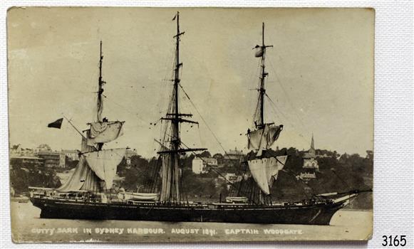 Three-masted ship in water, in front of a hill with buildings on it. 