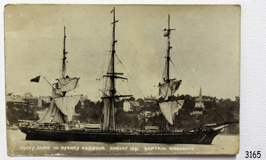 Three-masted ship in water, in front of a hill with buildings on it. 