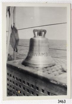 Ship's bell on horizontal surface, inscribed letters around bell, sea and sky as background.