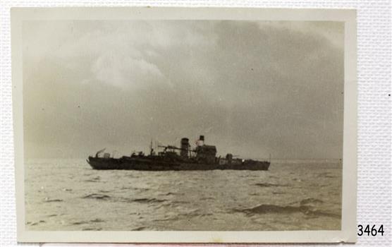 Ship is at sea, dark in contrast with its background of sea and sky.