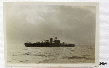 Ship is at sea, dark in contrast with its background of sea and sky.