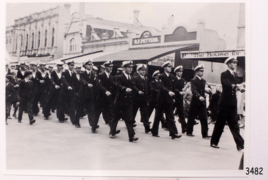Crew marching along a street in front of commercial buildings, with people watching